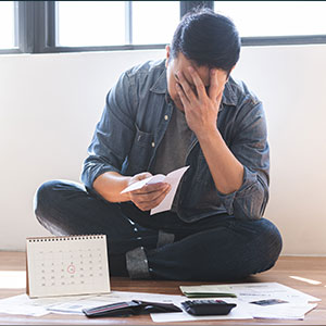 Man sitting on the floor with head in hands, looking at bills and a calendar, indicating financial stress.