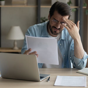 Distressed man reviews documents at a laptop, possibly due to bankruptcy fears.