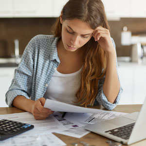 Worried young woman reviews documents with a calculator and laptop, suggesting financial concerns.